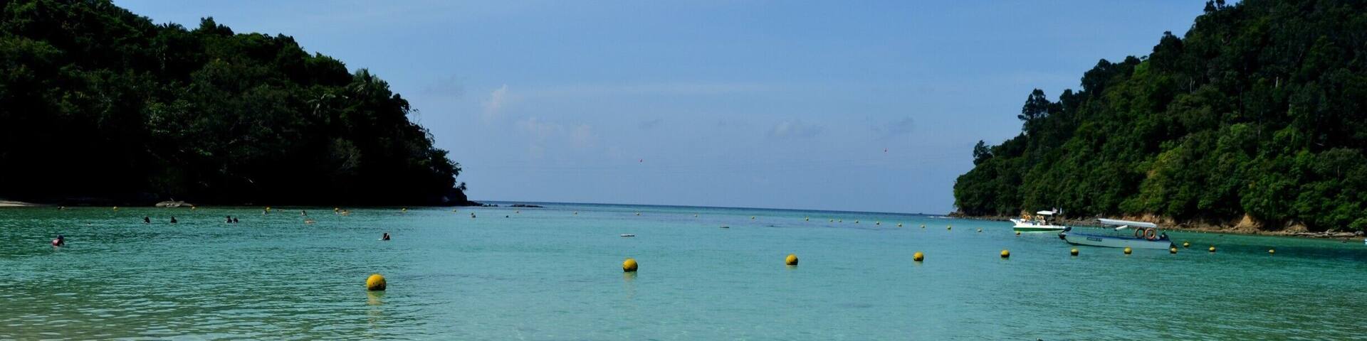 A clear turquoise sea with a sandy beach and lush green hills in the background.