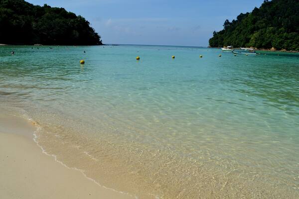 A clear turquoise sea with a sandy beach and lush green hills in the background.