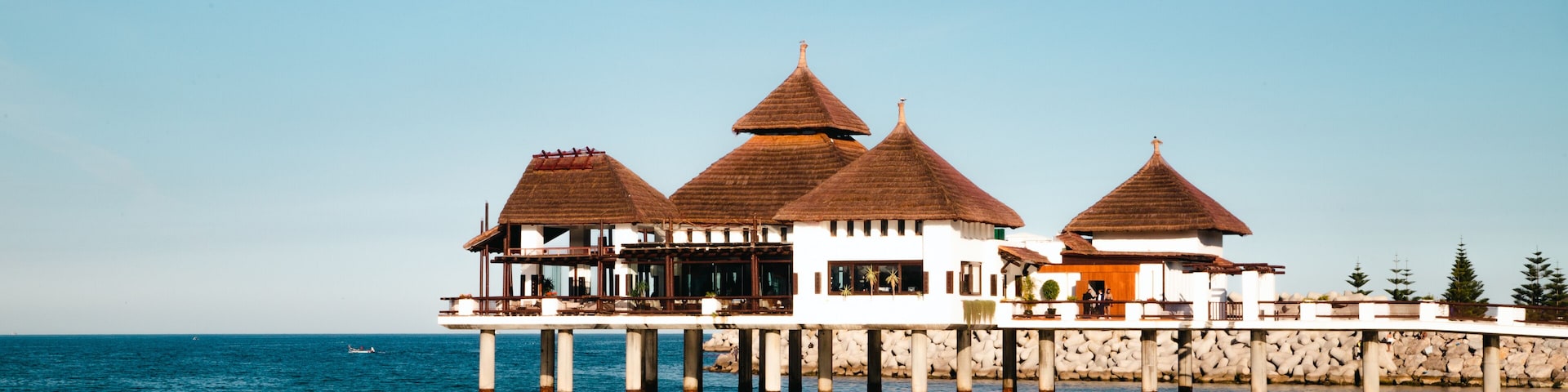 An elegant stilted structure with thatched roofs extends over the blue waters of M'diq, surrounded by a wooden walkway and greenery.