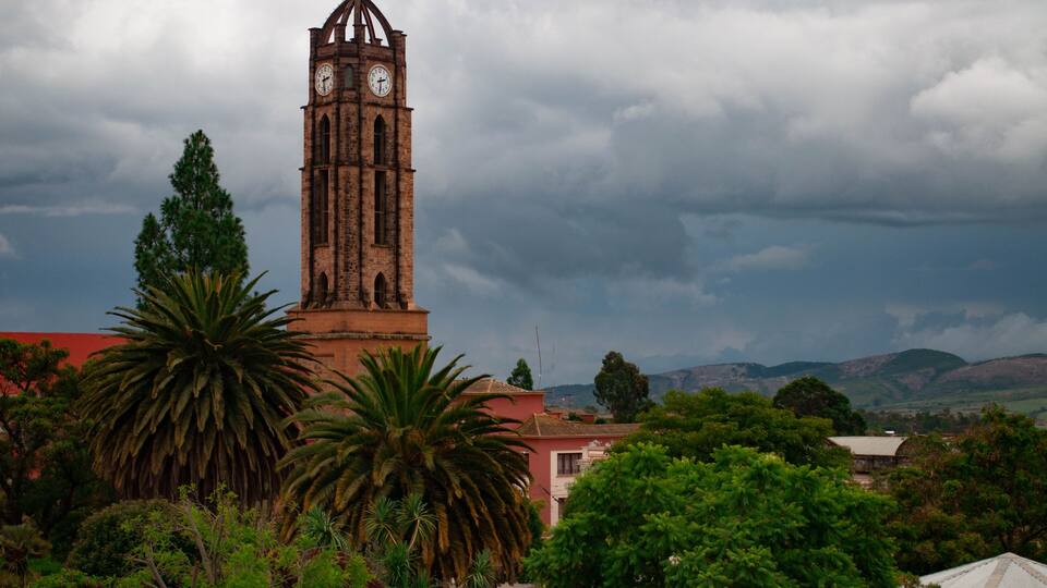 A tall clock tower with a cross on top, surrounded by lush greenery and a cloudy sky.