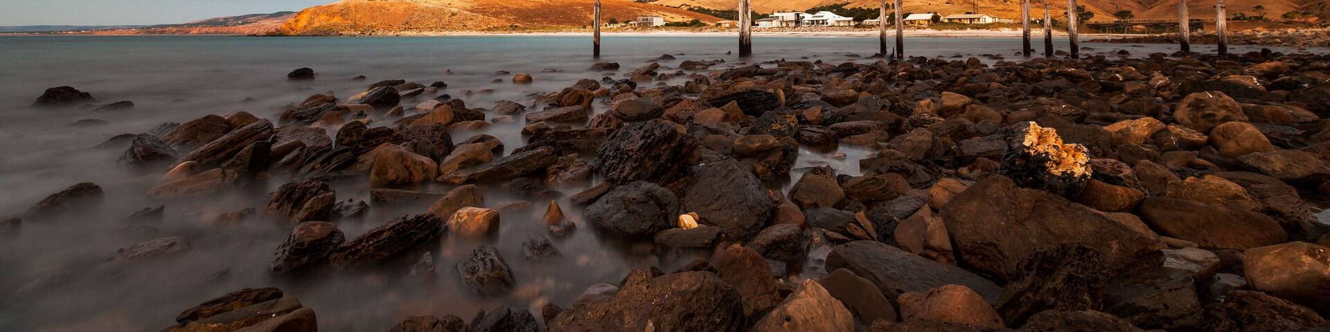 A rocky beach with a calm sea, wooden posts, and a distant village.