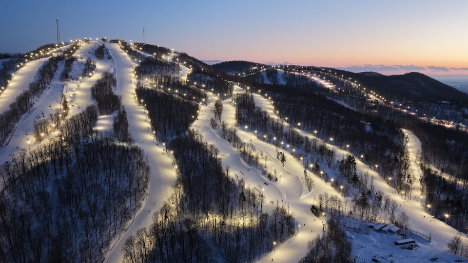 A snow-covered ski slope at dusk with illuminated paths and trees.