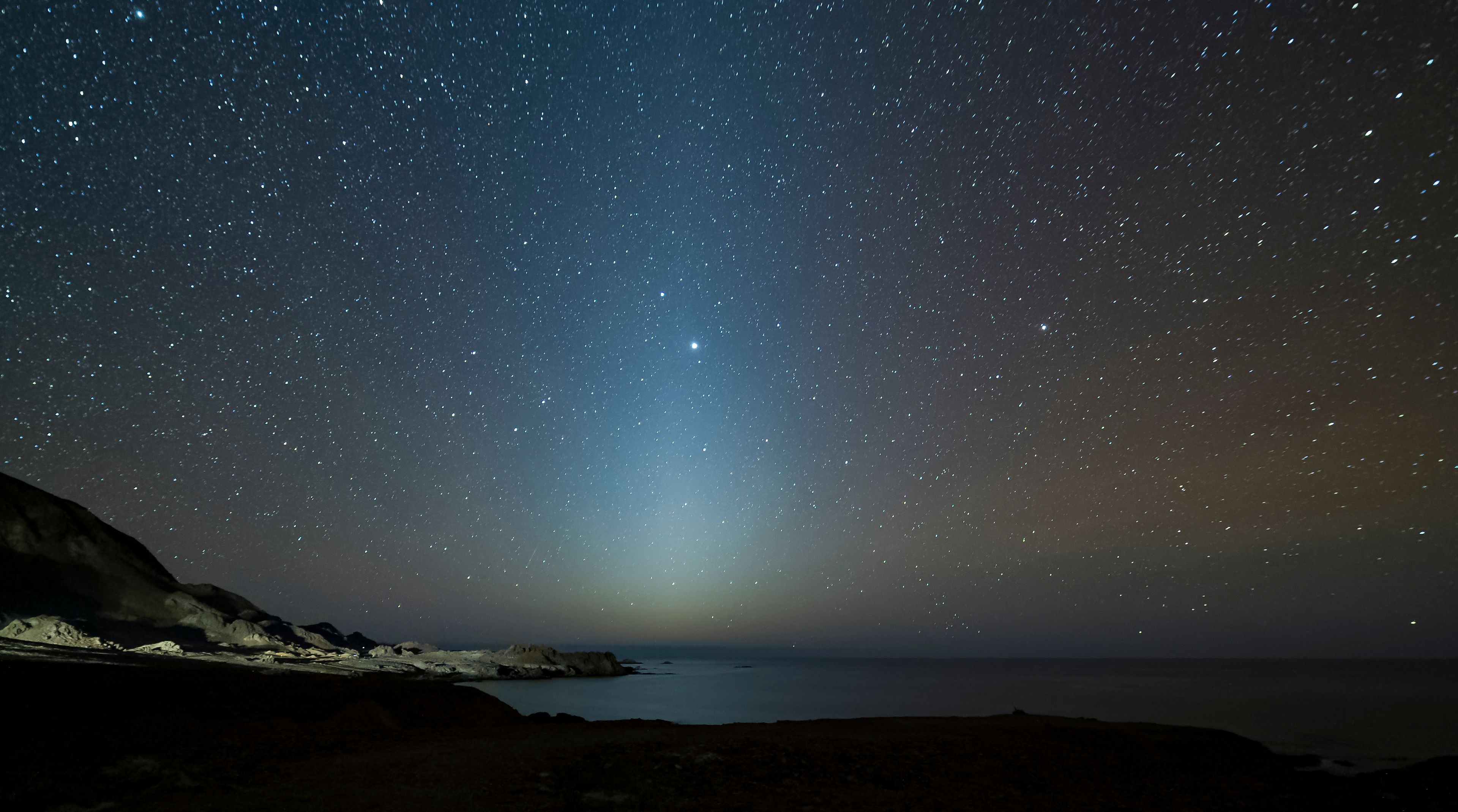 Cielo estrellado sobre un paisaje costero con una cadena montañosa visible.