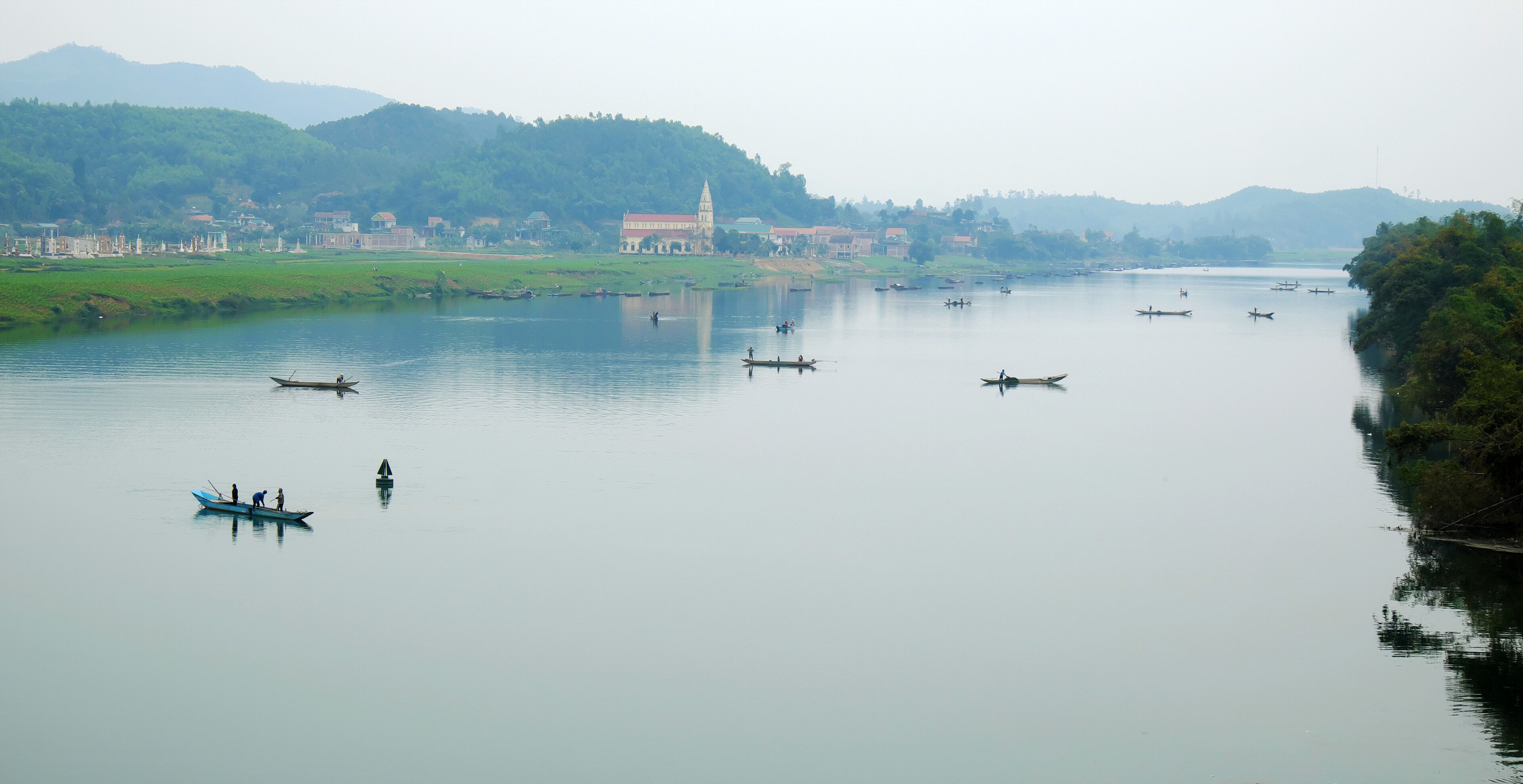 A calm river with several boats, a temple, and a forested hillside.