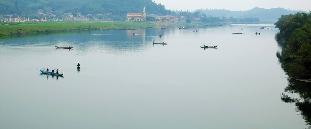 A calm river with several boats, a temple, and a forested hillside.