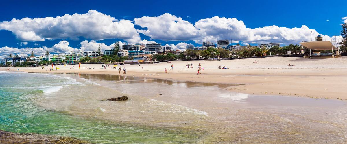 A beach with clear turquoise water, a rocky shoreline, and a cityscape in the background.