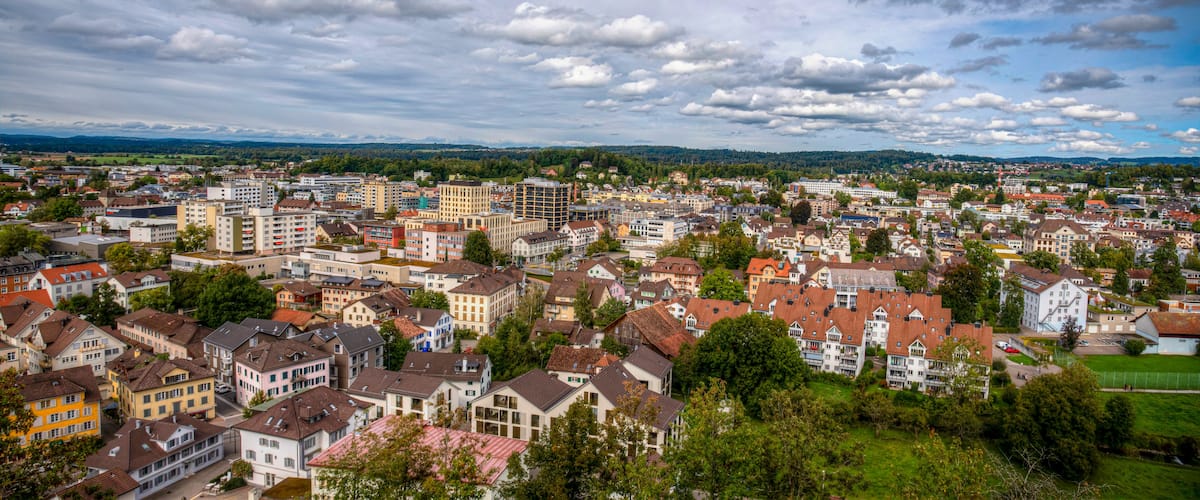 Une vue panoramique d’une ville où se côtoient des immeubles résidentiels et, au loin, un paysage vallonné sous un ciel partiellement nuageux.