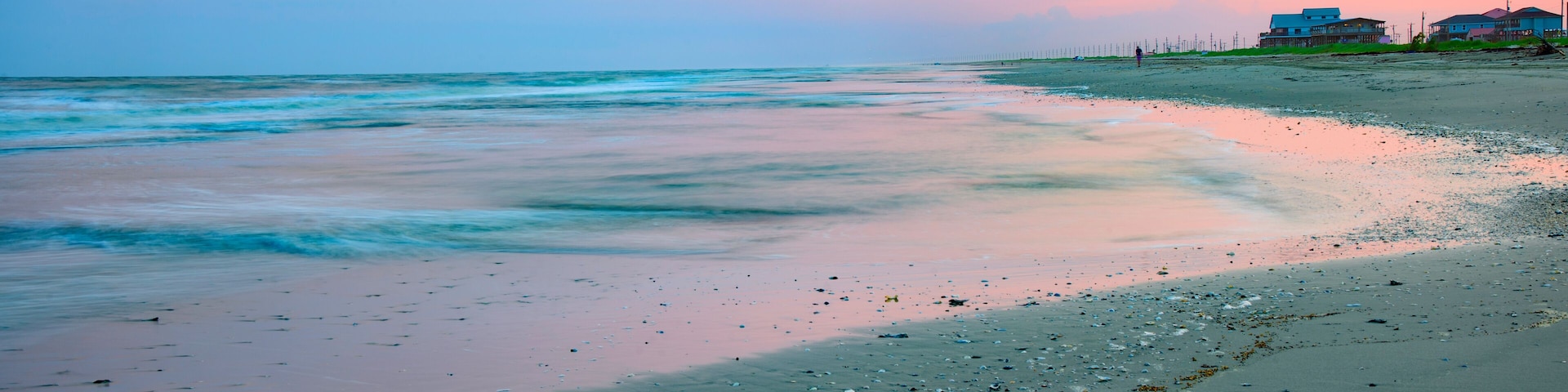 Ein Strand mit sanften Wellen, blauem Himmel und Häusern im Hintergrund.