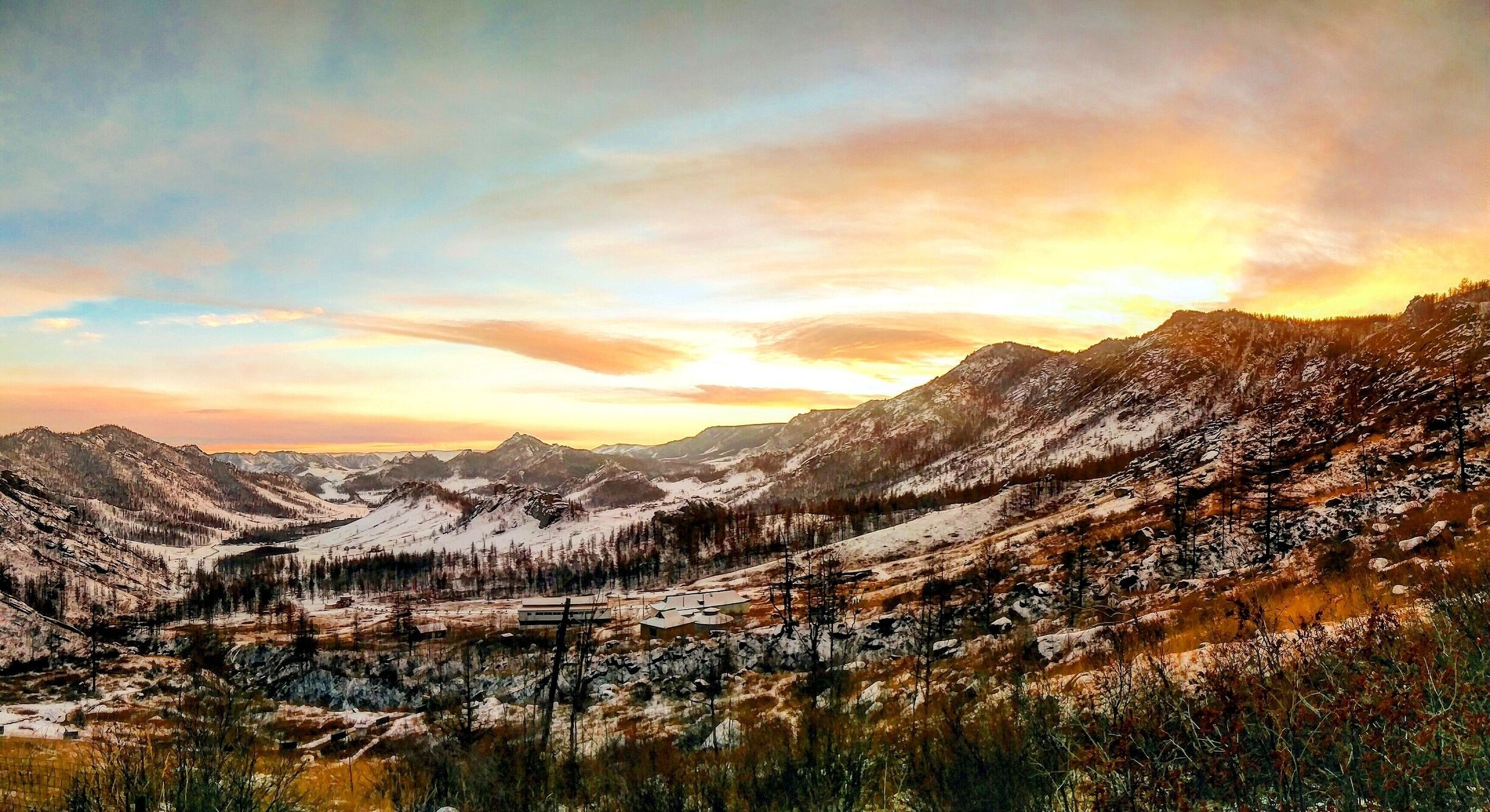 A mountainous landscape with snow-covered peaks, a valley, and a small settlement with buildings.