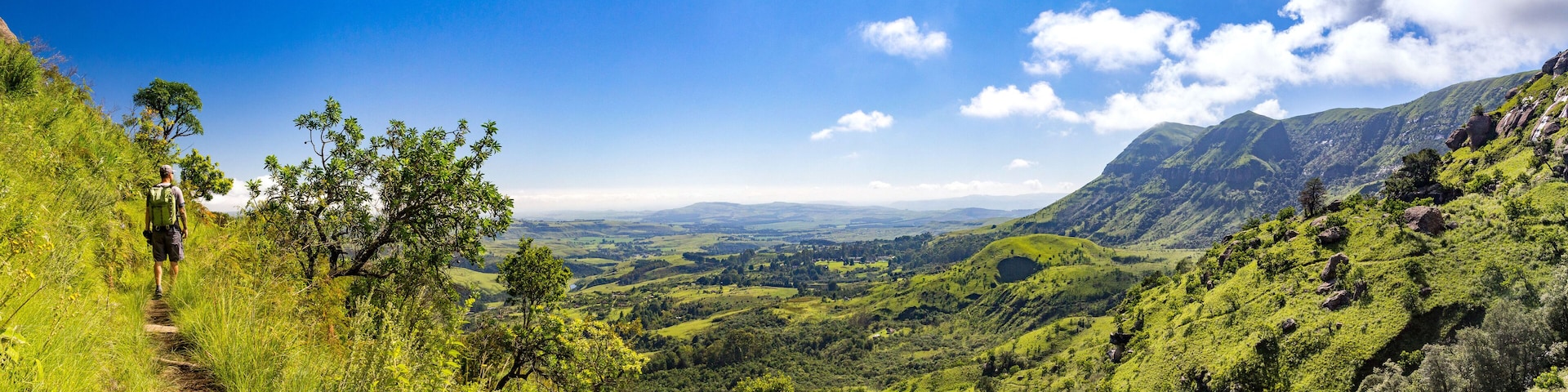 Man hiking on a small trail with a panoramic view over steep and green mountains on a sunny day, Drakensberg, Giants Castle Game Reserve, South Africa