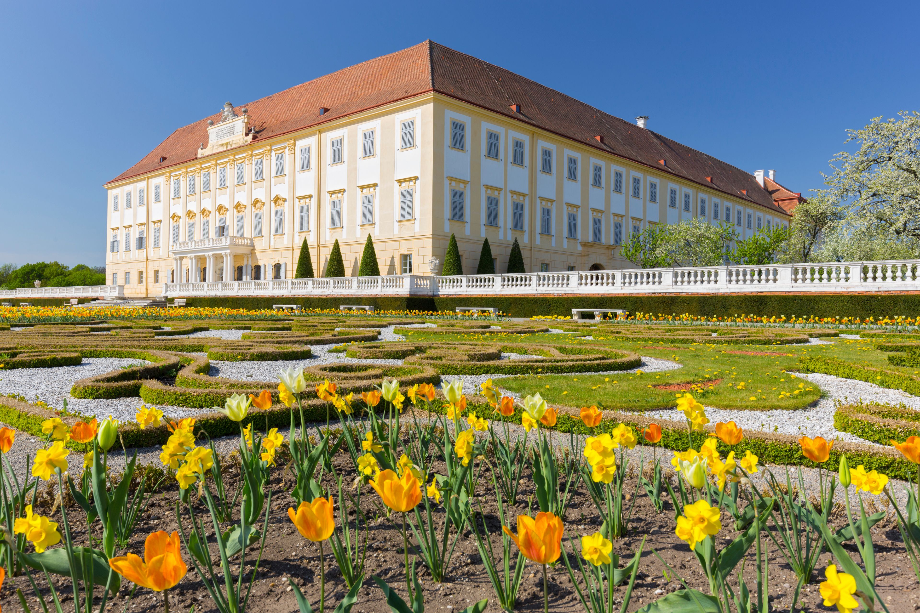Ein großzügiges, klassizistisches Gebäude mit einer symmetrischen Fassade, ein gepflegter Garten mit geometrisch angelegten Blumenbeeten und ein klarer, blauer Himmel.