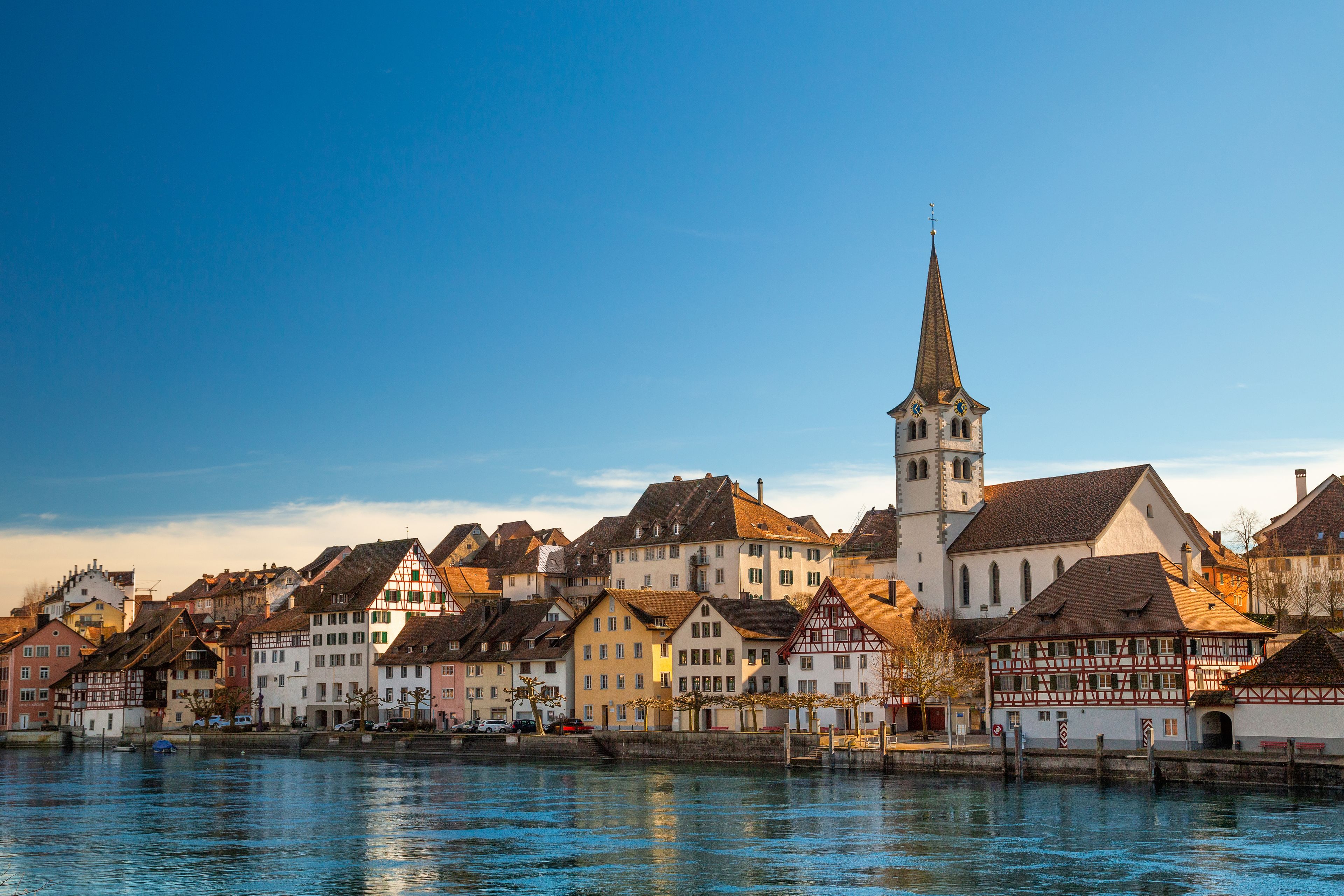 A riverside town with historic buildings, a prominent church tower, and a clear blue sky.