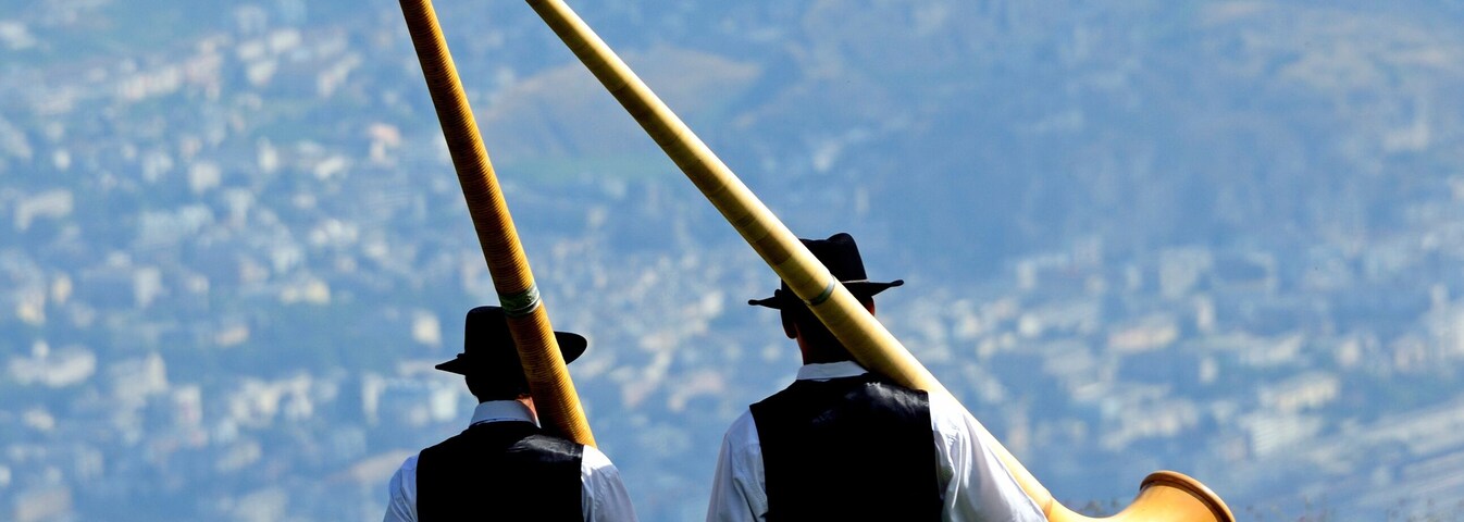 Nendaz, Conthey District,
Valais, Wallis, Switzerland, Europe - two men in traditional black vests, trousers and hats walking with alphorn on their arms, Swiss Alps, Nendaz village seen in valley