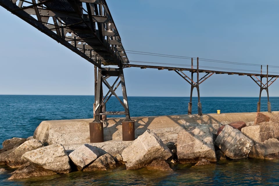 A red lighthouse on a rocky pier with a metal bridge overhead.