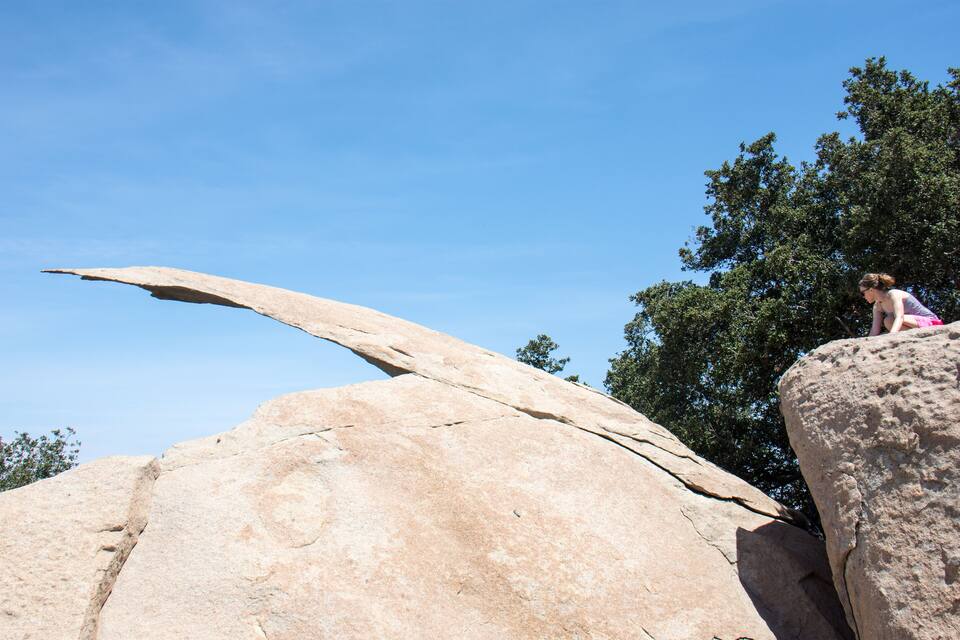 A large rock formation with a prominent overhang and a person sitting on a smaller rock nearby.