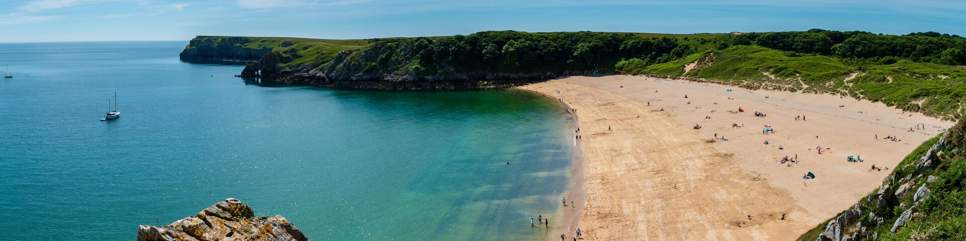 Uma praia costeira com águas turquesa cristalinas, areal arenoso e um falcão rochoso em primeiro plano.