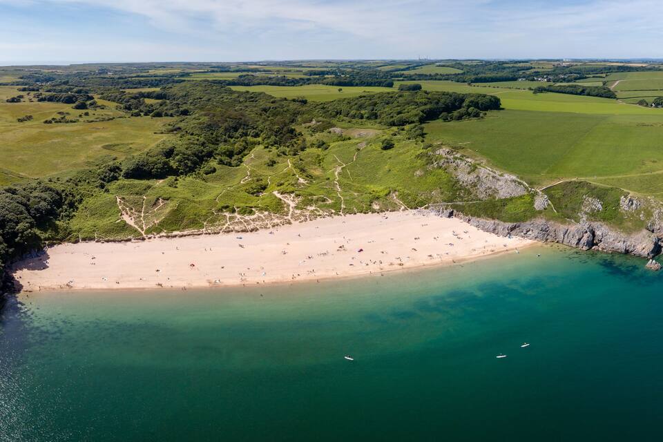 Un paysage côtier avec une plage de sable, des eaux turquoise et limpides, et des collines verdoyantes en arrière-plan.