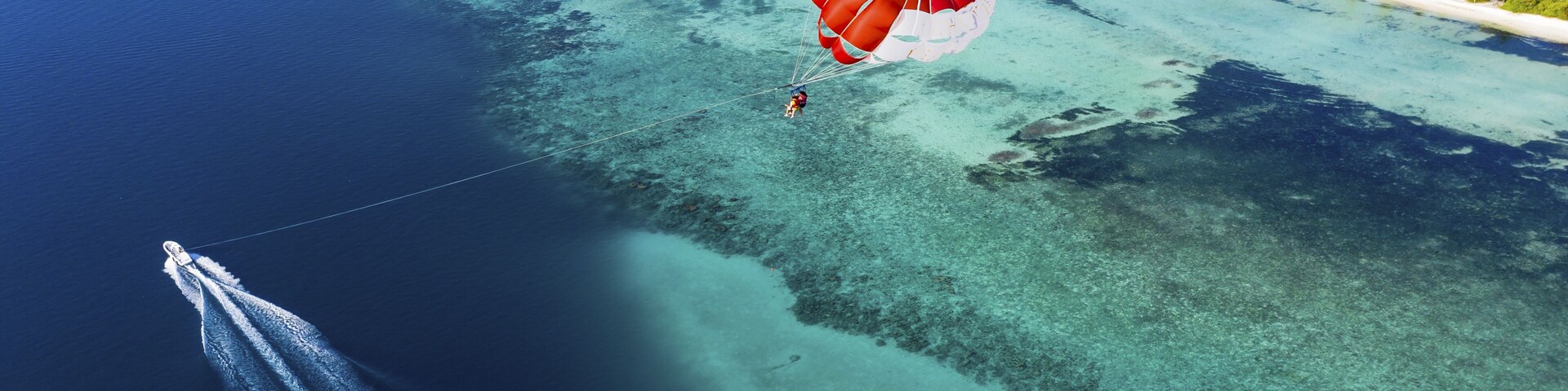 Uma pessoa praticando parapente sobre um mar azul-turquesa, com vista clara para uma ilha tropical e um cais.