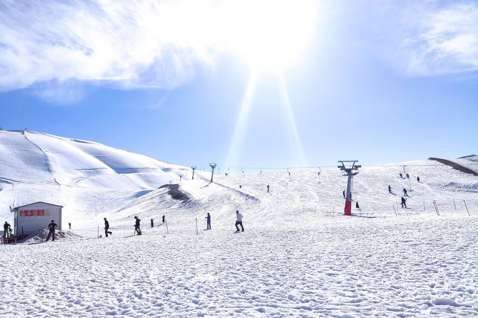 Pista de esqui coberta de neve, com esquiadores, um teleférico e um pequeno edifício.