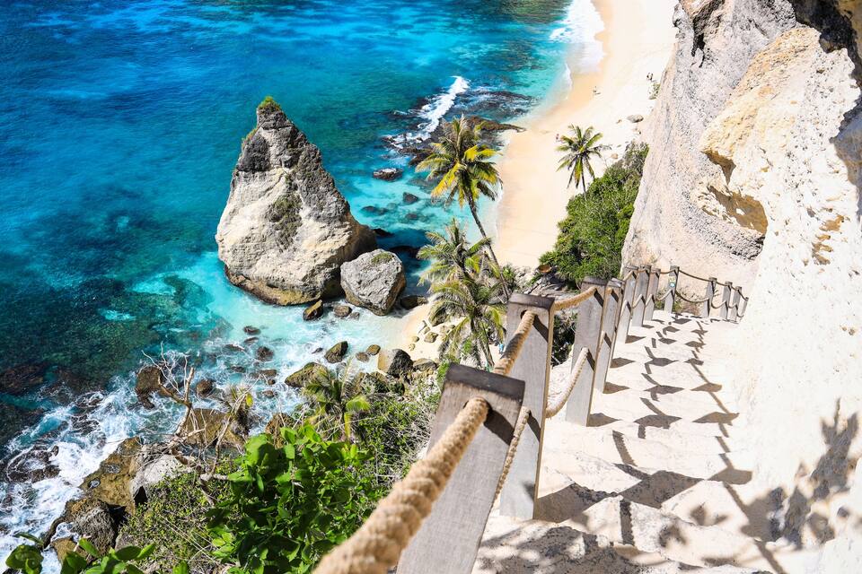 A coastal landscape with clear turquoise waters, a prominent rock formation, a sandy beach, and a staircase leading down to the shore.