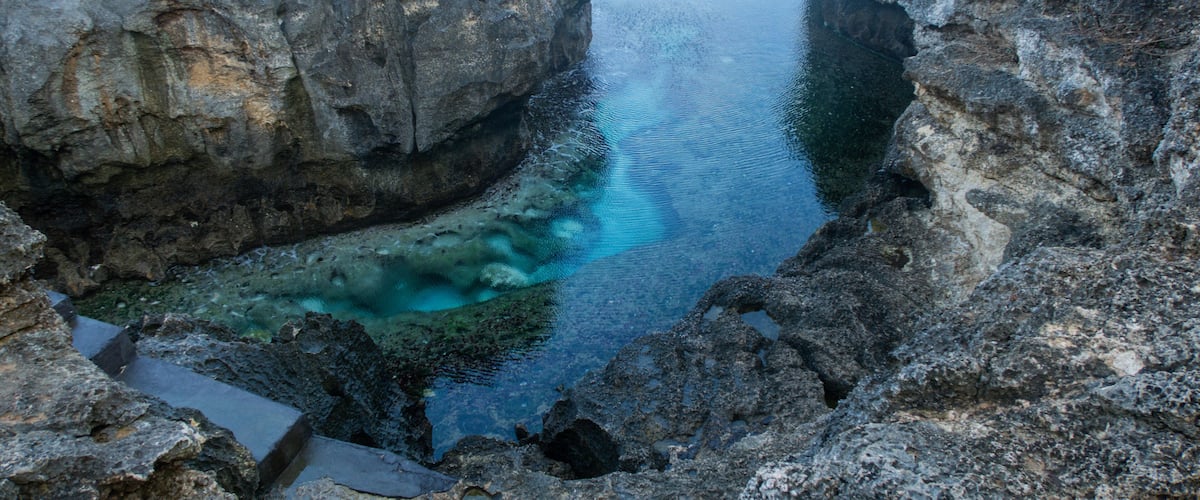 A narrow water channel between rocky cliffs with clear water reflecting the sky.