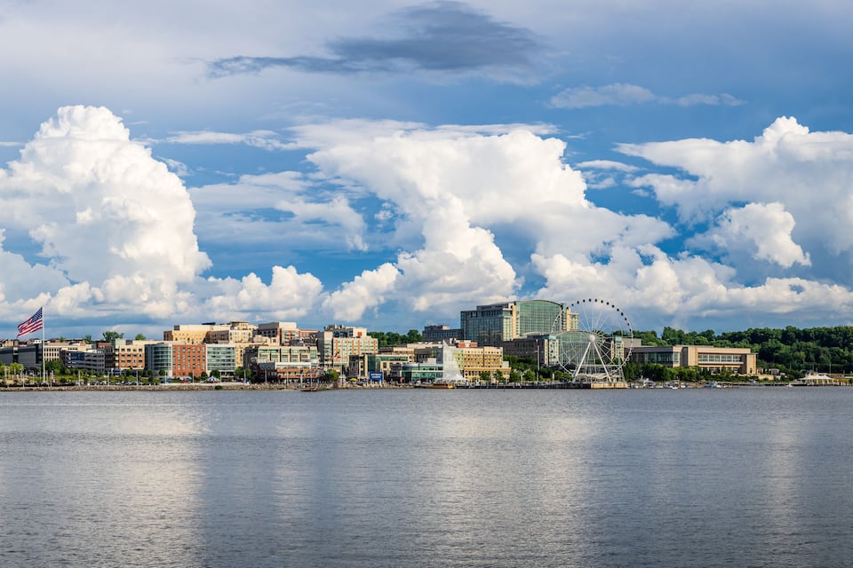 A waterfront cityscape with a prominent Ferris wheel, a large American flag, and a mix of modern and historic buildings.