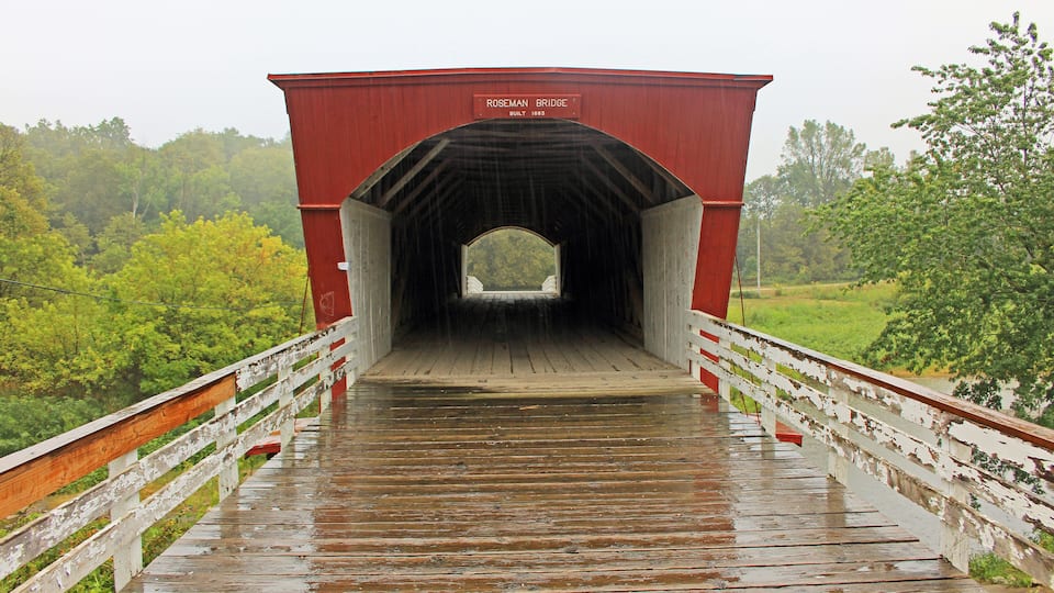 A red covered bridge with a sign reading "Roseman Bridge Built 1883" in a forested area.