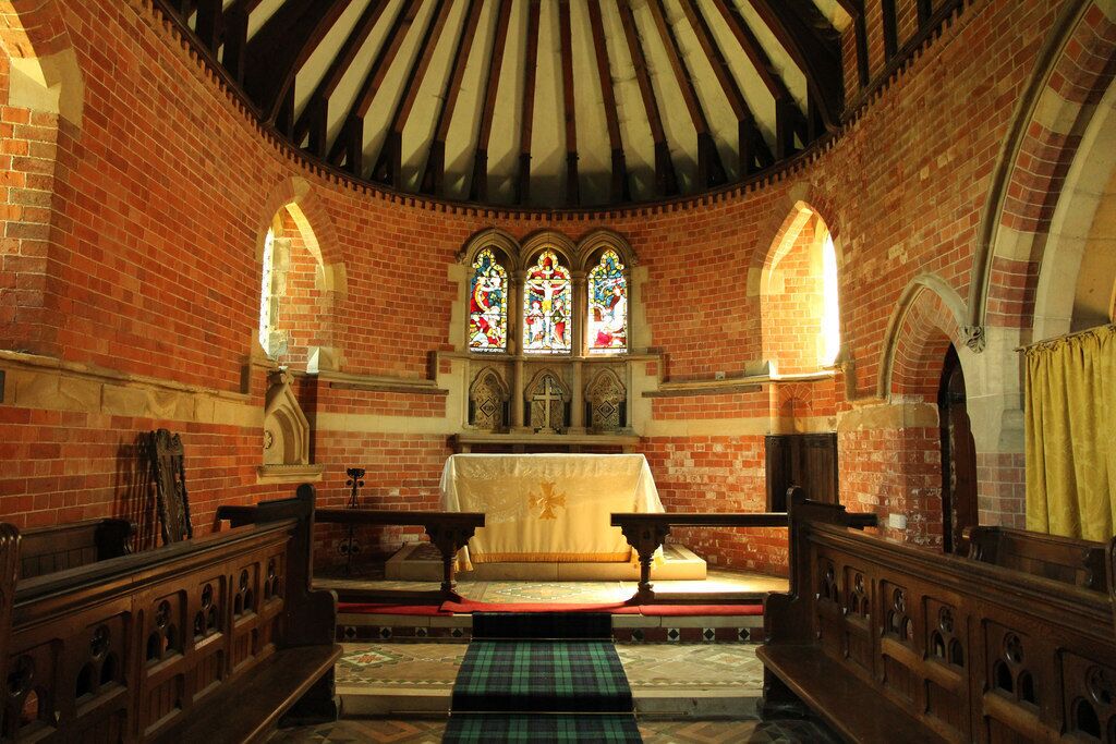 A church interior with brick walls, wooden pews, a central altar, and stained glass windows.