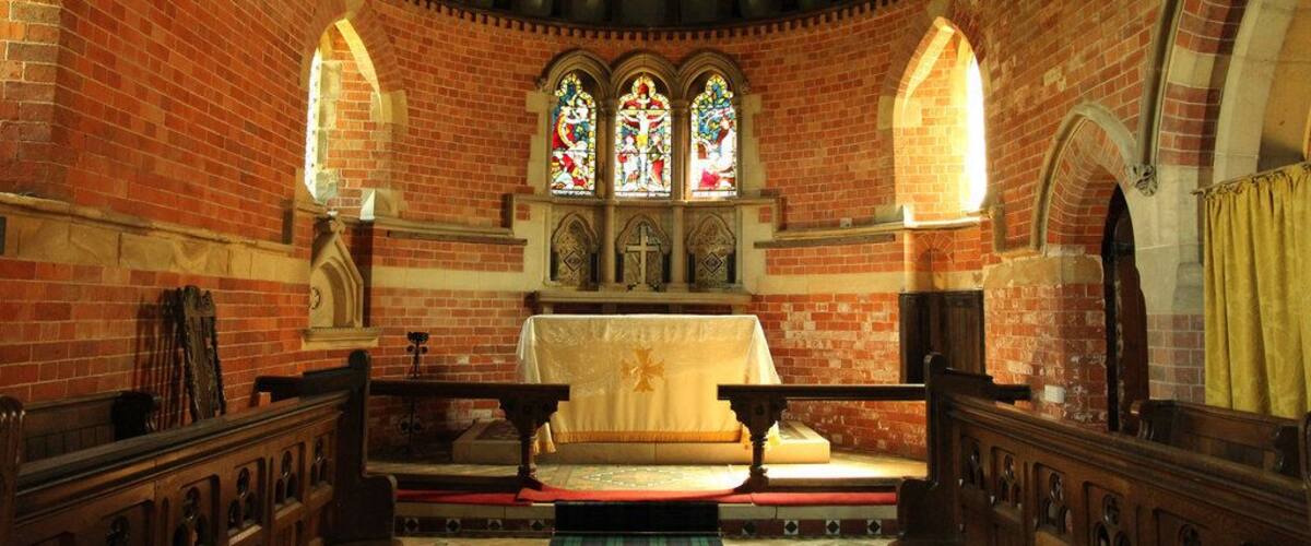 A church interior with brick walls, wooden pews, a central altar, and stained glass windows.