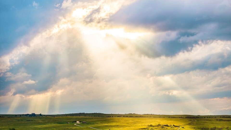 A vast green field with a winding path, under a dramatic sky with rays of sunlight piercing through clouds.