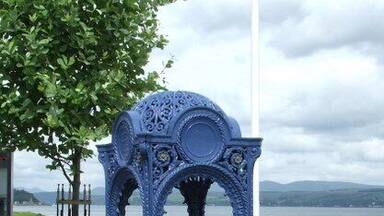 A blue gazebo with intricate designs, surrounded by grass and a body of water in the background.