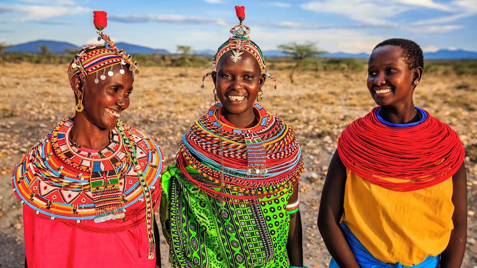 Three individuals in traditional attire with colorful patterns and accessories, standing in a dry, open landscape with sparse vegetation and distant mountains.