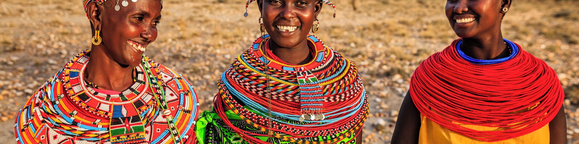 Três pessoas vestindo roupas típicas com padrões e acessórios coloridos, em uma paisagem seca e aberta, com vegetação rala e montanhas ao longe.