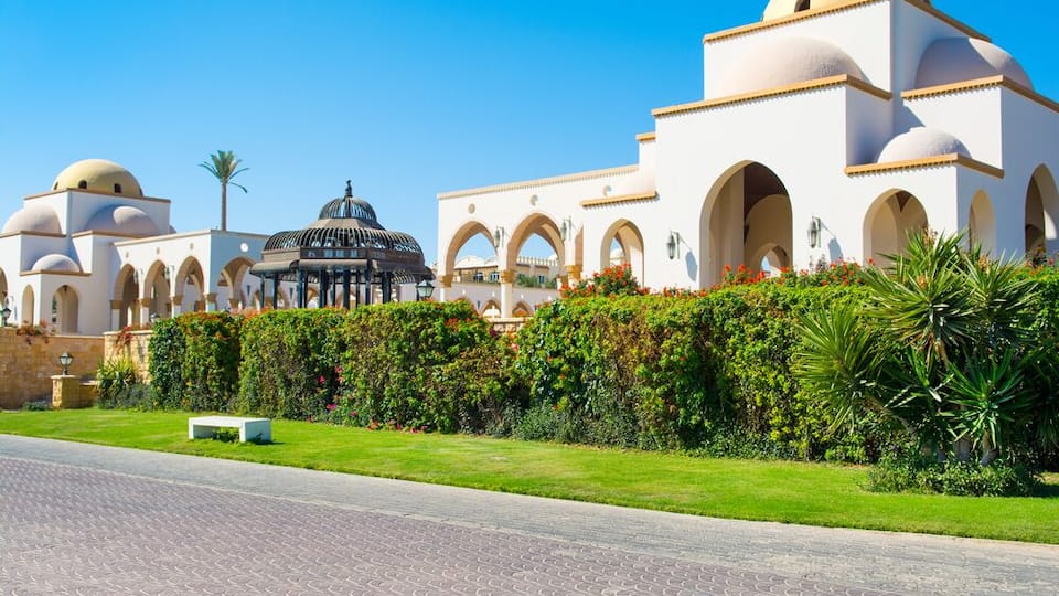 A large white building with arches and domes, surrounded by well-maintained gardens and a paved road.