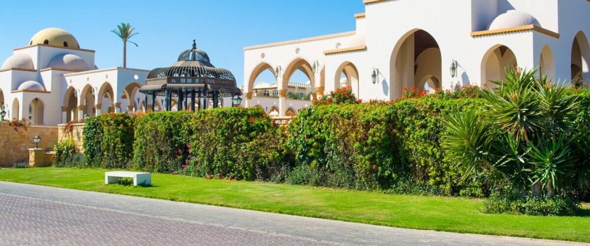 A large white building with arches and domes, surrounded by well-maintained gardens and a paved road.