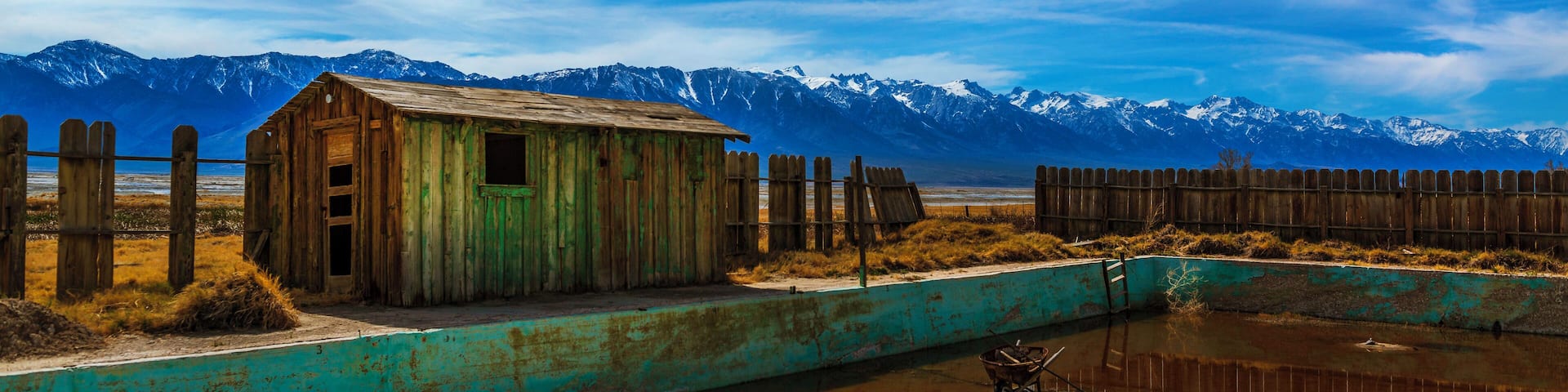 A rustic wooden shack by a turquoise pond with mountains in the background.