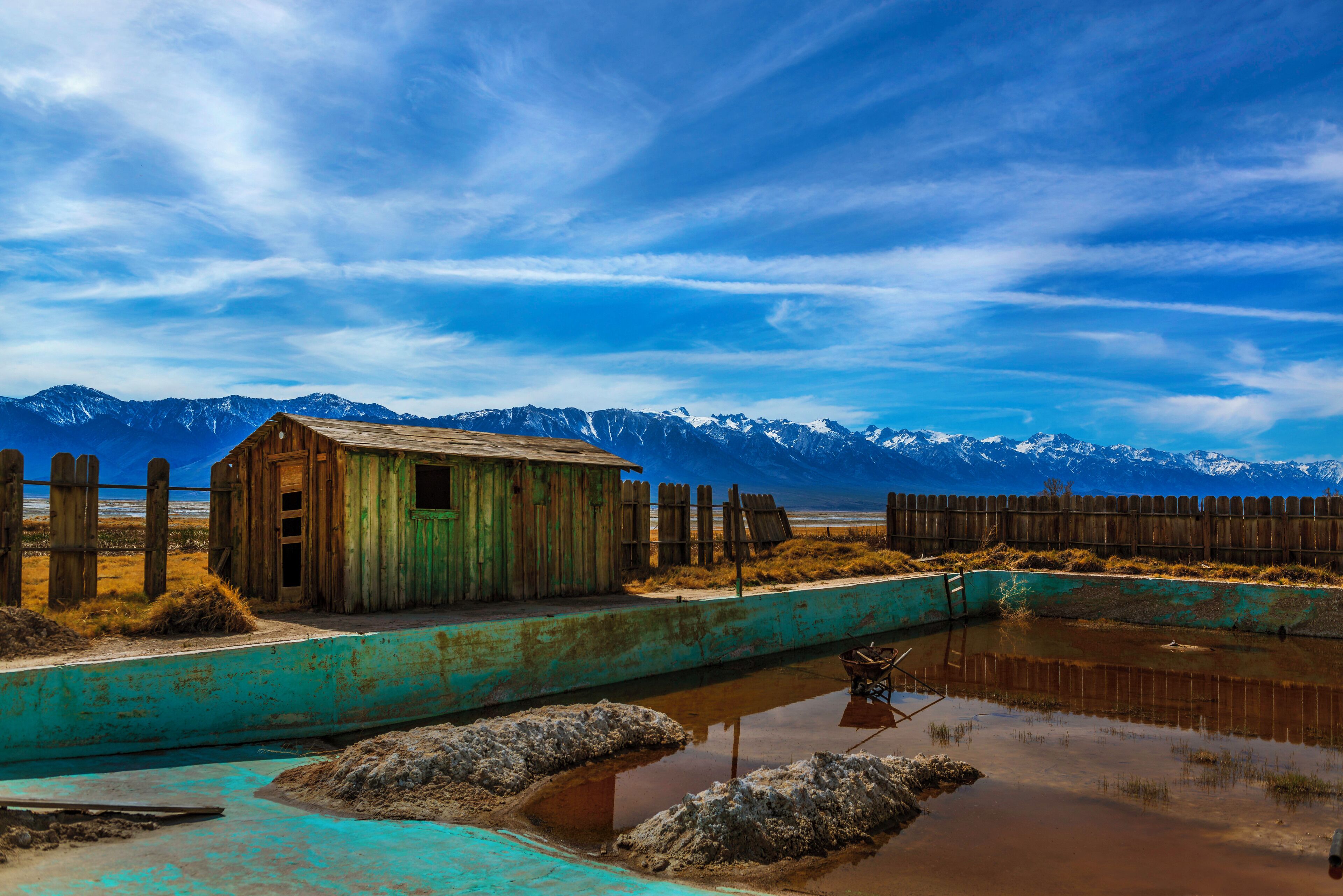 A rustic wooden shack by a turquoise pond with mountains in the background.