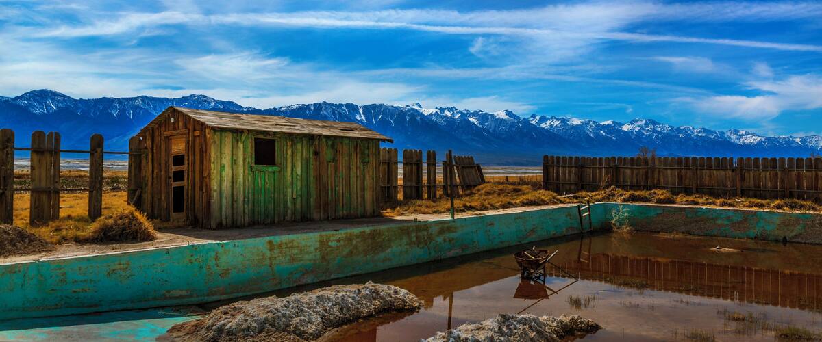 A rustic wooden shack by a turquoise pond with mountains in the background.
