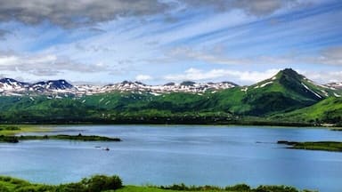 A lake surrounded by green hills and mountains under a partly cloudy sky.