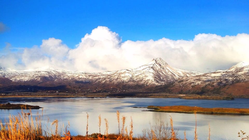 A snow-capped mountain range, a calm lake, and autumn grasses in the foreground.
