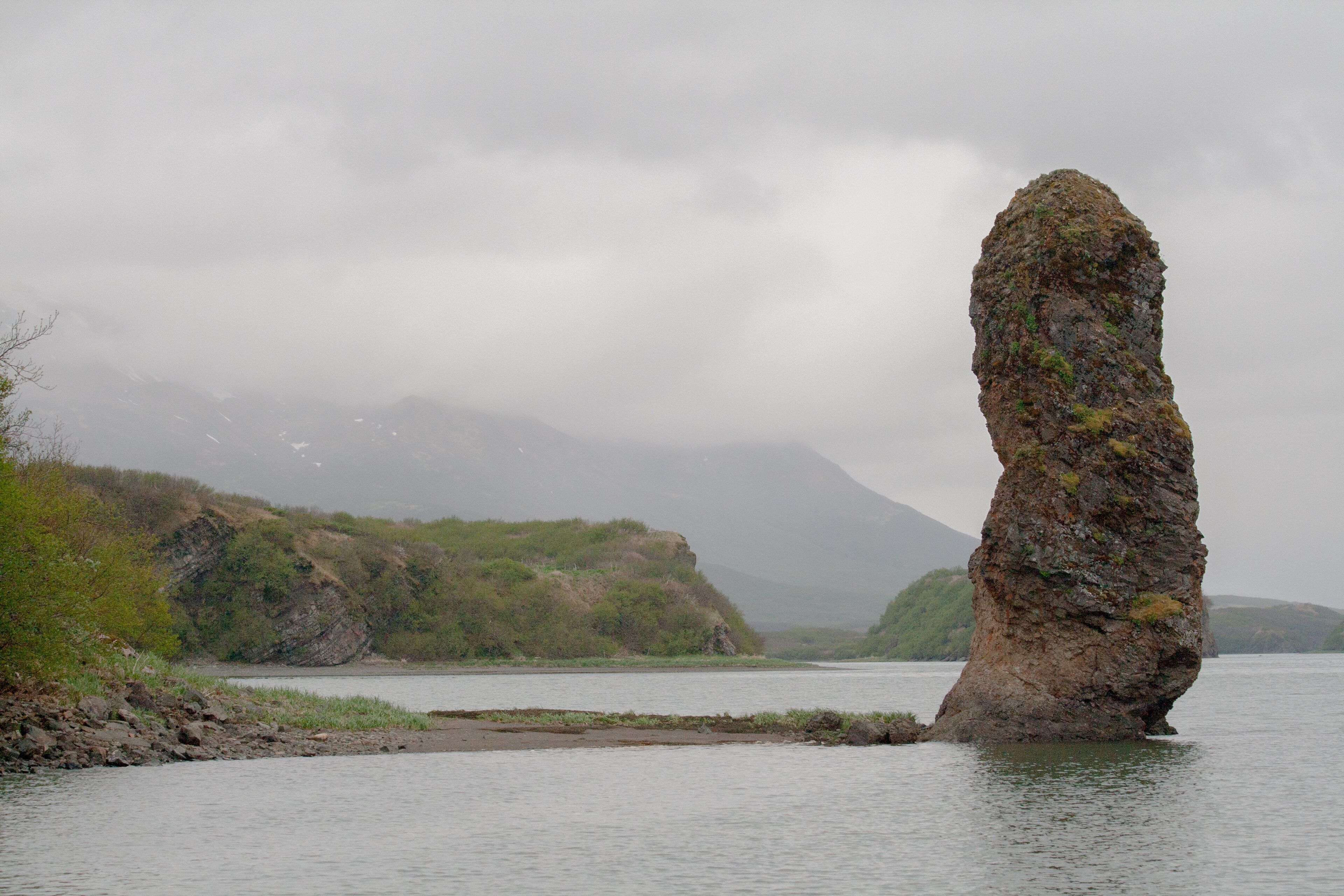 A solitary rock formation in a body of water, surrounded by hills and a cloudy sky.