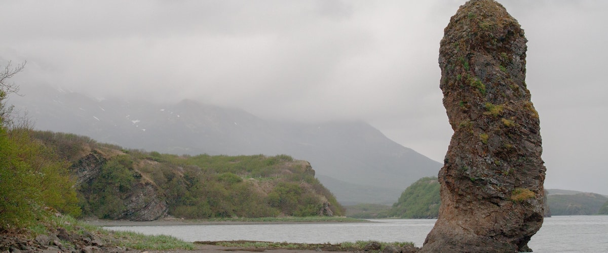 A solitary rock formation in a body of water, surrounded by hills and a cloudy sky.