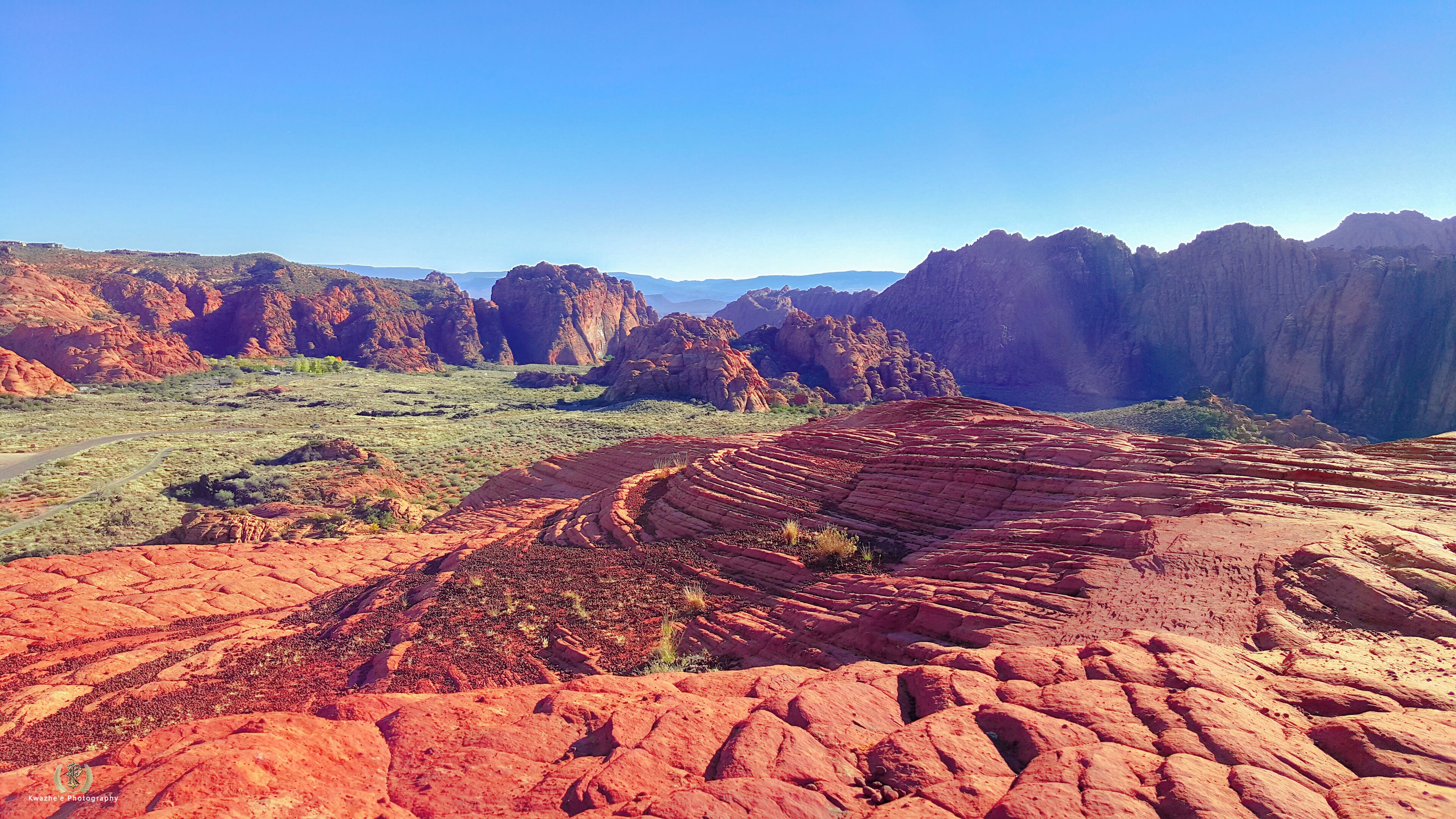 A vast desert landscape with prominent red rock formations and a clear blue sky.