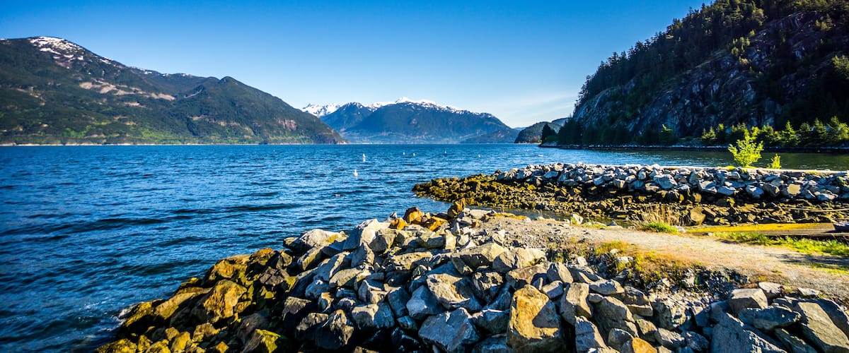 A rocky shoreline with a clear blue lake, mountains in the background, and a forested hill.