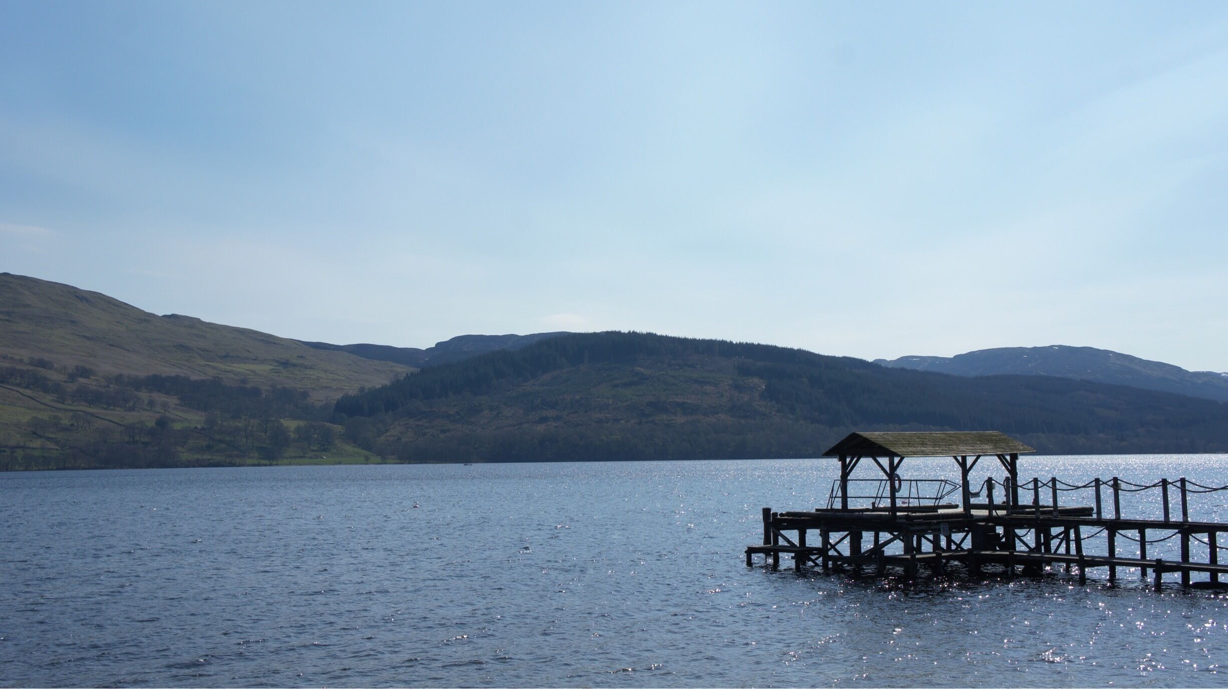 A wooden pier extending into a calm lake, surrounded by rolling hills and a clear sky.