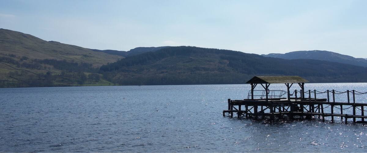 A wooden pier extending into a calm lake, surrounded by rolling hills and a clear sky.