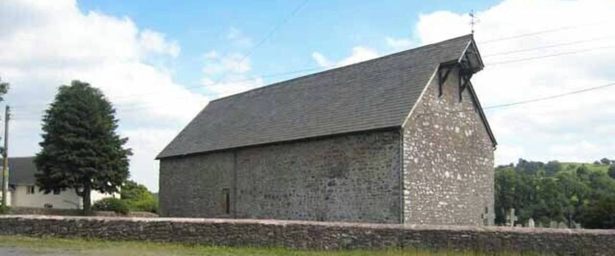 A large stone building with a sloped roof, surrounded by a stone wall and a grassy field.