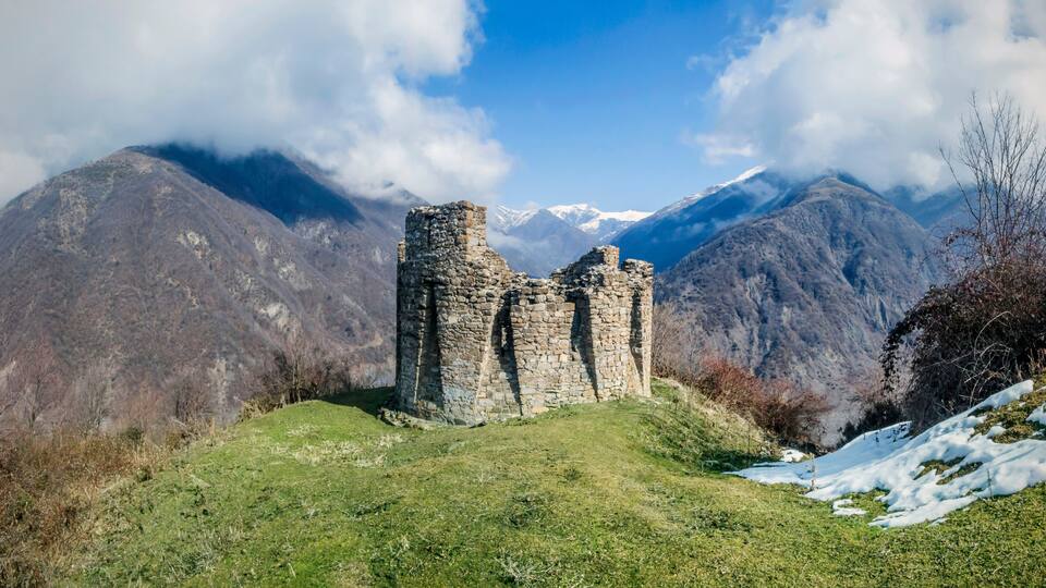 A stone ruin on a grassy hill with snow patches, surrounded by mountains and a partly cloudy sky.