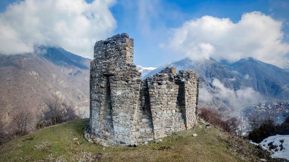 A stone ruin with two towers, surrounded by mountains and a village in the valley.