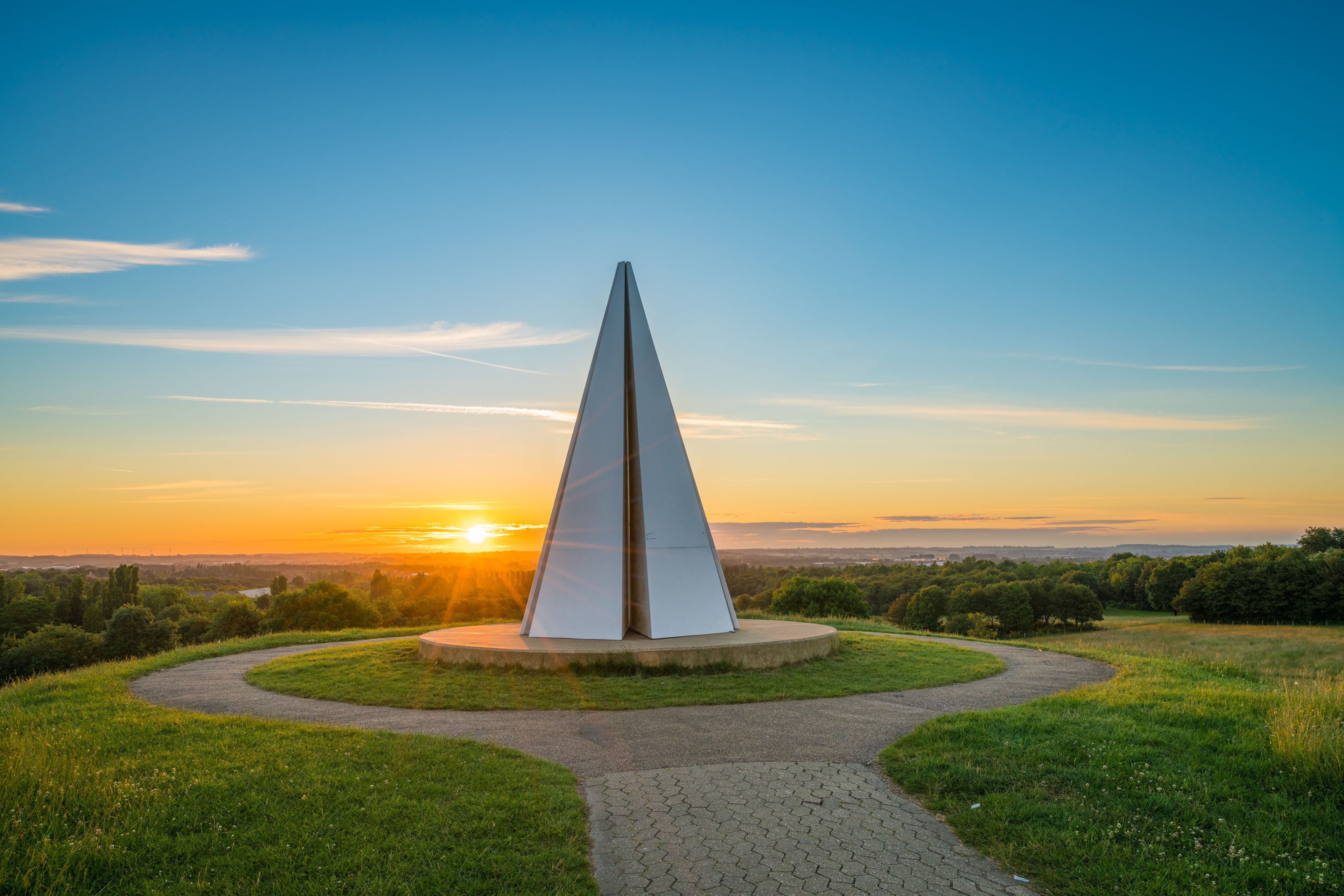 A tall, slender monument with a pointed top, surrounded by a circular pathway and grassy areas, set against a backdrop of a sunset sky.